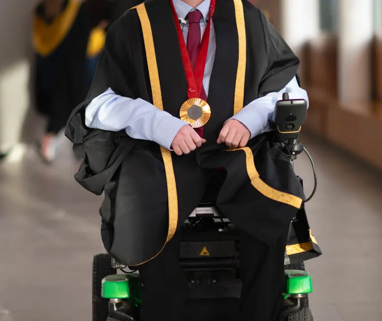Stephen McGuire, a Paralympic gold medallist, wearing formal graduation attire with gold trim and a medal around his neck, seated in a wheelchair. He is smiling and positioned in a well-lit hallway, with blurred figures of graduates in the background.