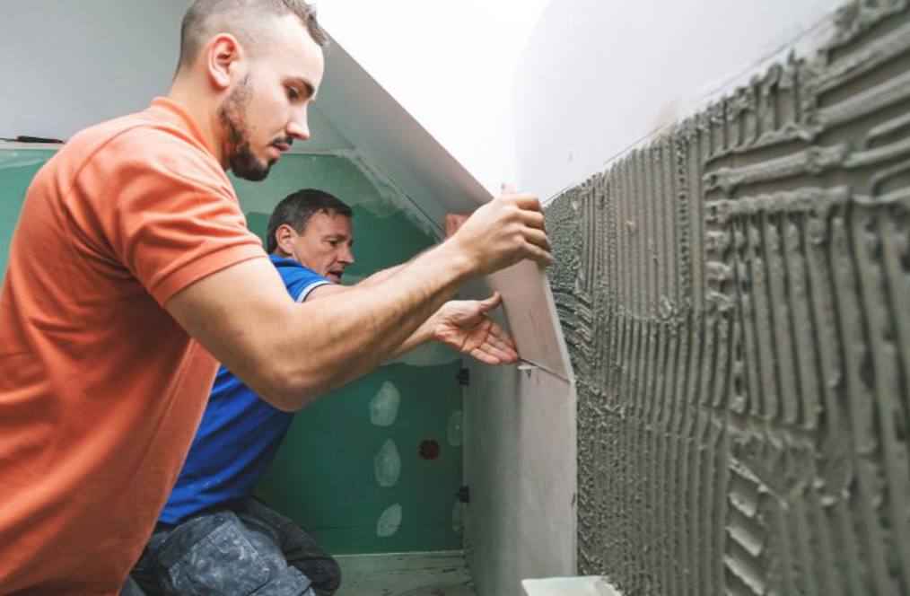 Two men tiling a wall in a room under construction. The man in the foreground is applying adhesive to the wall with a notched trowel, while the man in the background prepares to assist, highlighting teamwork in a hands-on construction or trade setting.